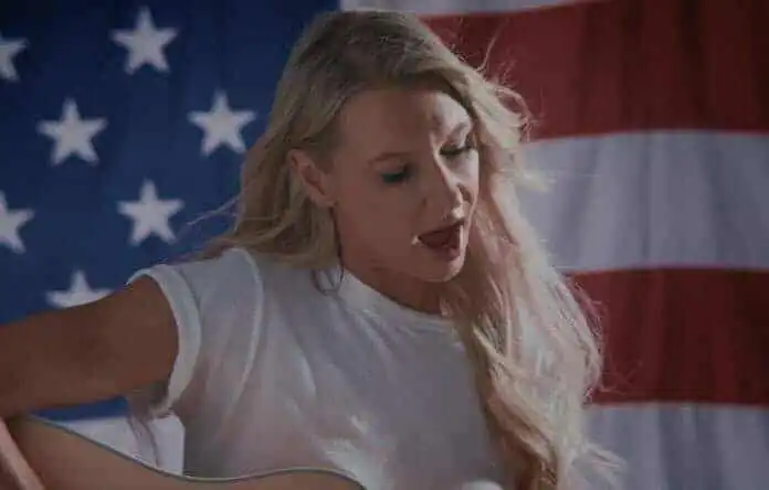 A woman with long blonde hair singing and playing guitar against an American flag backdrop.