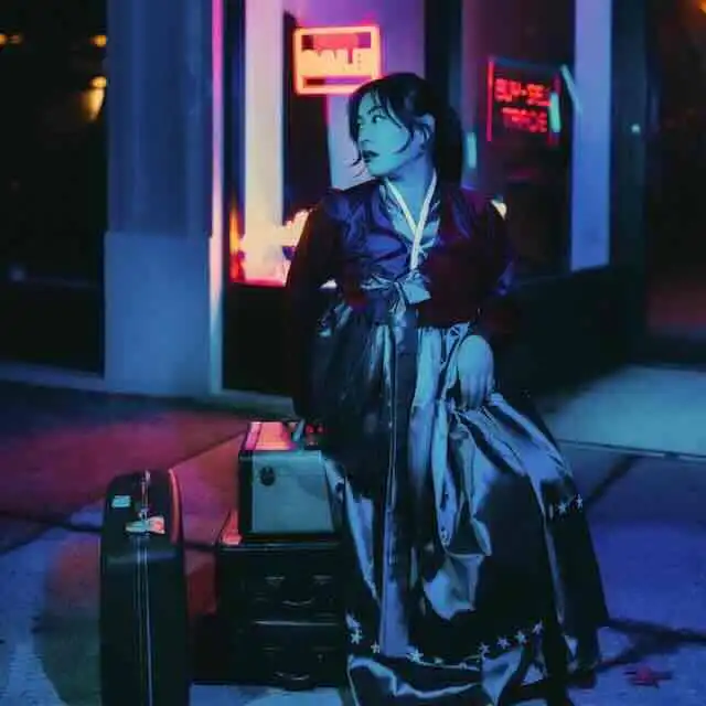 Teen traveler with luggage at night outside a neon-lit building.