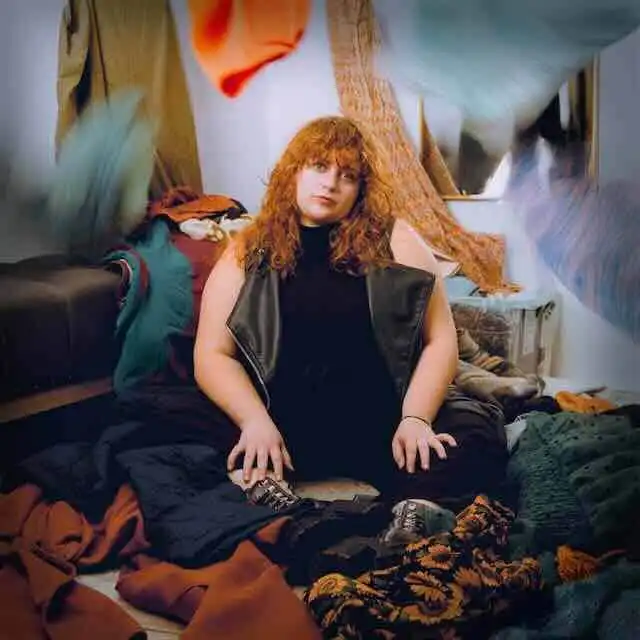 Woman sitting on the floor surrounded by clothes and fabric decor, showcasing confidence and style.