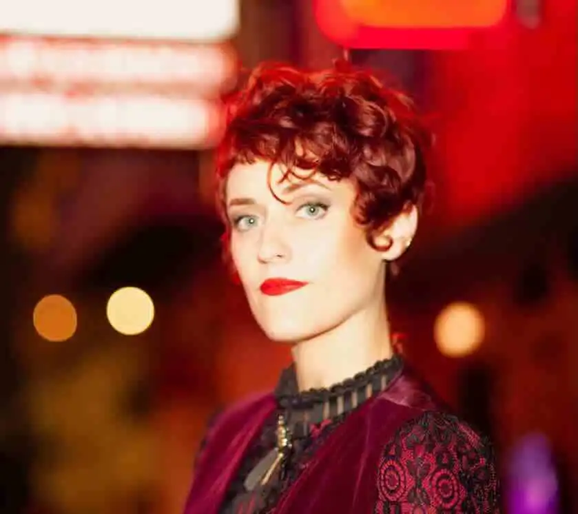 Redhead woman with short curly hair and red lipstick in a vibrant nightlife setting.