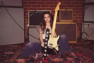 Young woman holding an electric guitar in a music rehearsal space with amplifiers and brick wall background.