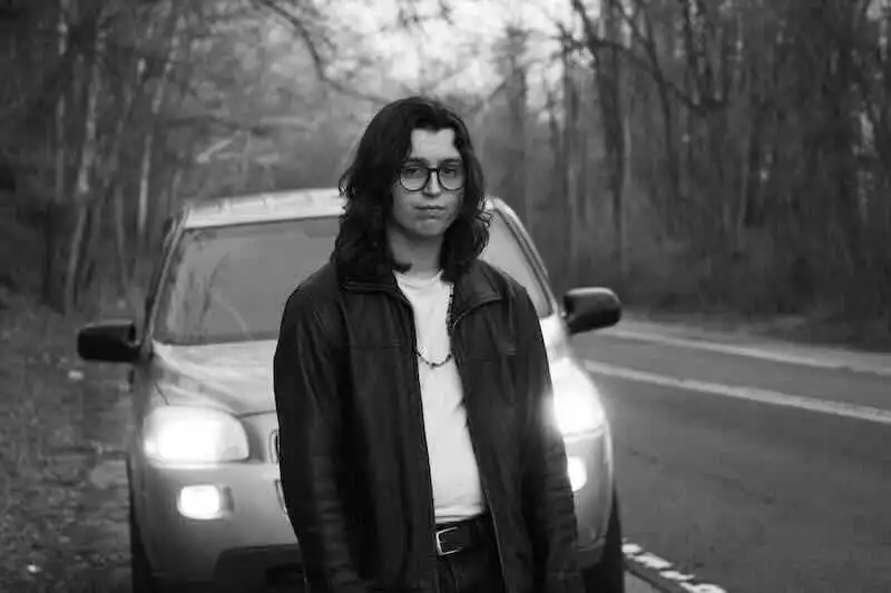 A young person with long dark hair and glasses standing in front of a car on a wooded road in black and white.