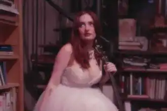 A woman with red hair in a white dress holds a guitar while sitting among bookshelves in a warm, intimate library environment.