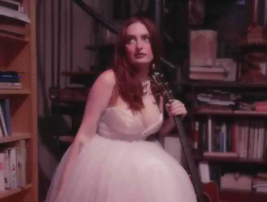 A woman with red hair in a white dress holds a guitar while sitting among bookshelves in a warm, intimate library environment.