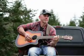 A man sitting on a truck bed playing acoustic guitar in a natural outdoor setting with trees.
