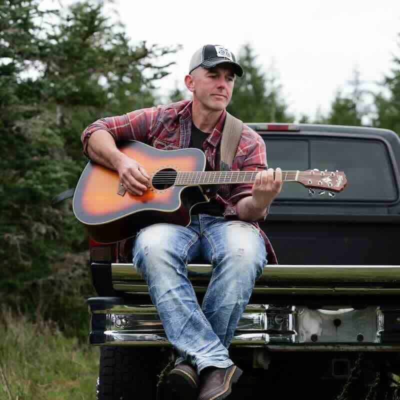 A man sitting on a truck bed playing acoustic guitar in a natural outdoor setting with trees.