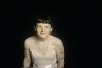 Portrait of a smiling woman with tattoos in a lace dress against a black background.