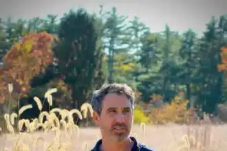 A man in a navy polo shirt stands in a golden field with autumn trees in the background, capturing peace and natural beauty.