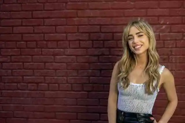 Smiling blonde woman in casual outfit standing against red brick wall, showcasing confidence and positivity.