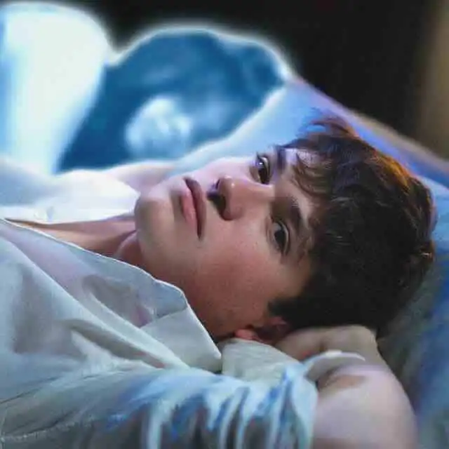 Close-up of a young man lying on his side in bed, looking thoughtful and relaxed.