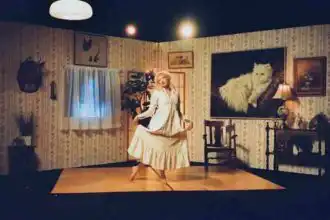 A woman in vintage dress poses in a retro-style living room with floral wallpaper and cozy decor.