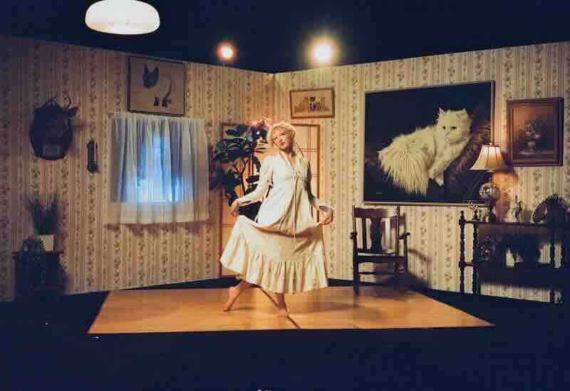 A woman in vintage dress poses in a retro-style living room with floral wallpaper and cozy decor.