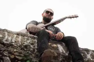 Man with guitar sitting on stone wall, wearing sunglasses, outdoor setting.
