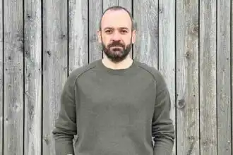 A man with a beard and casual gray sweater poses against a rustic wooden background.
