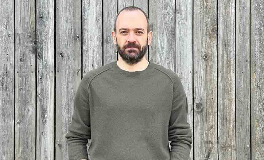 A man with a beard and casual gray sweater poses against a rustic wooden background.