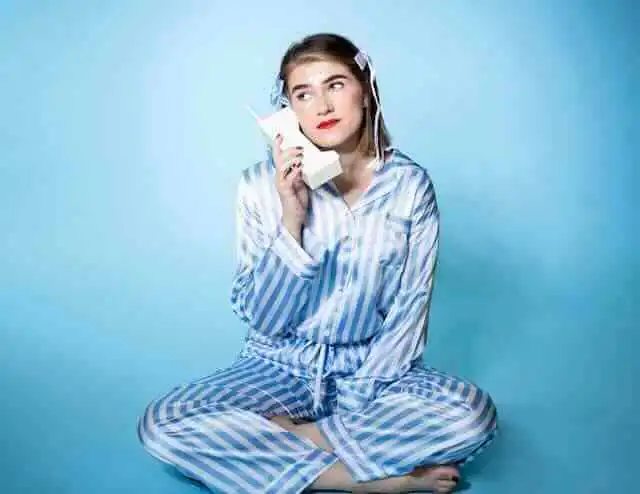 Young woman in pajamas talking on vintage phone against blue background.