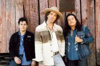 A stylish music band with three members posing outdoors against a rustic wooden background.