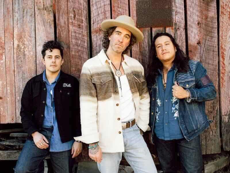 A stylish music band with three members posing outdoors against a rustic wooden background.