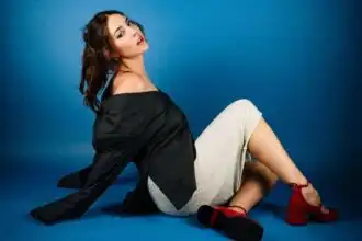 Stylish woman sitting against blue background, fashion-forward look, red heels, and confident expression.