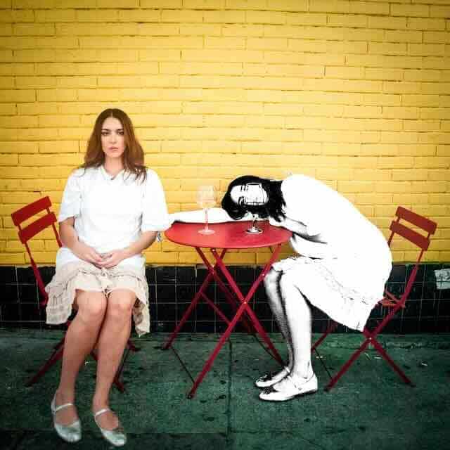 Two women sit at a small red table outside, one looking sad and the other resting her head on the table.