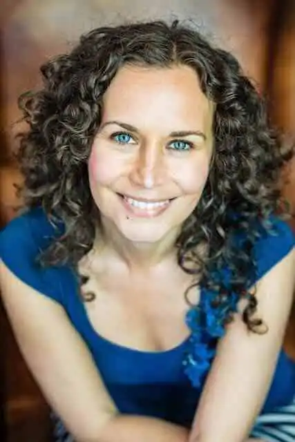 Close-up of a smiling woman with curly hair and blue eyes, wearing a blue top.