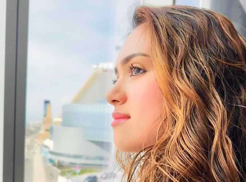 Beautiful woman looking out a window, urban background, natural lighting, thoughtful expression.