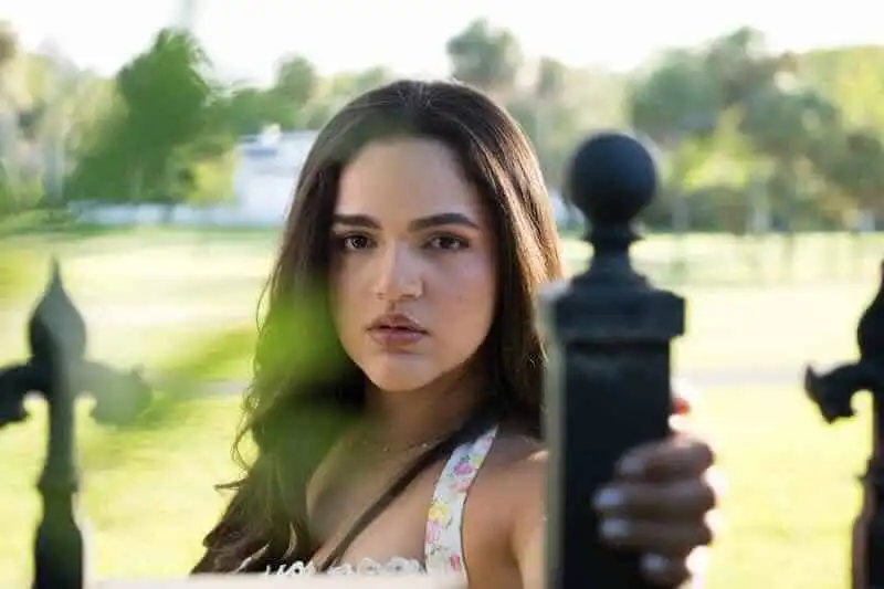 Close-up of a woman with long dark hair in a sunny outdoor setting, looking directly at the camera.
