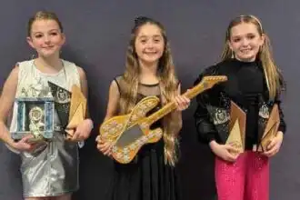 Three smiling young girls with awards and musical instruments at a celebration event.