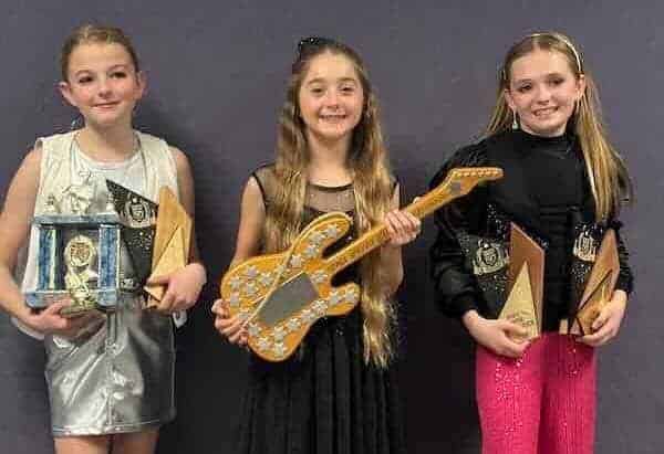 Three smiling young girls with awards and musical instruments at a celebration event.