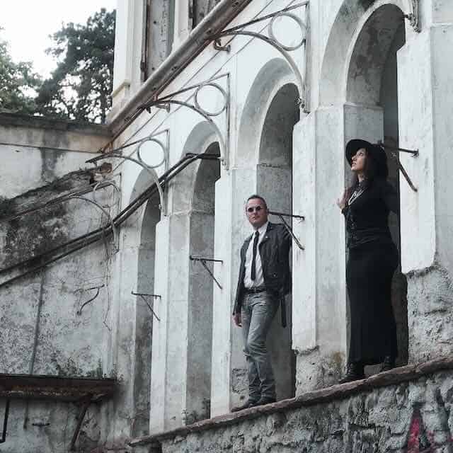 Stylish couple in black and gray outfits standing on ancient stone ledge near arched windows.