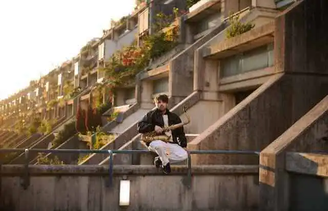 Man sitting on railing in front of hillside apartment buildings with modern architecture and greenery.