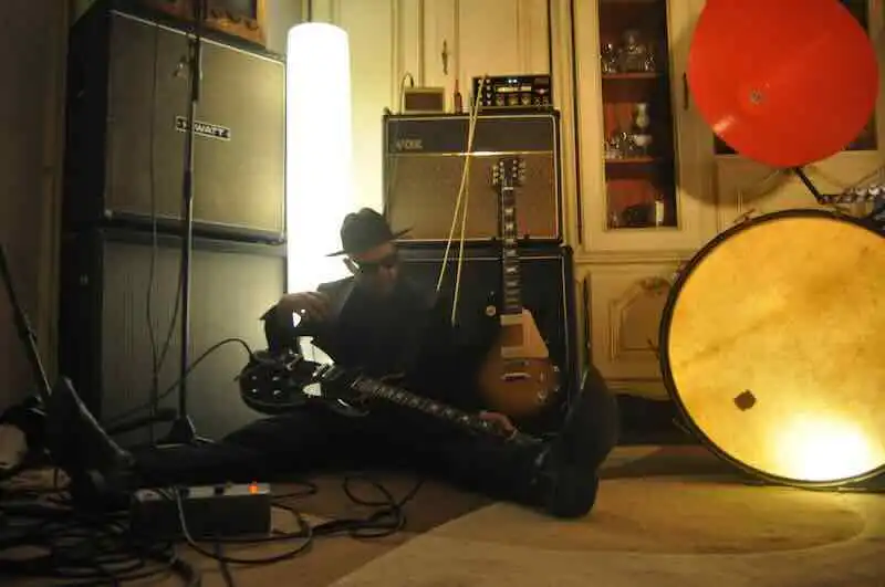Young man playing electric guitar in a cozy, vintage room with musical gear and warm lighting.