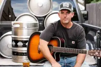 Man playing acoustic guitar outdoors with beer and beer kegs in background.
