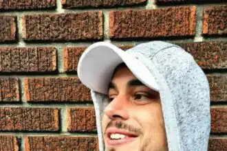 Close-up of a smiling man wearing a gray hoodie and cap outdoors with brick background.