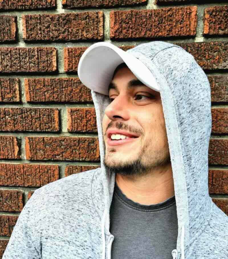 Close-up of a smiling man wearing a gray hoodie and cap outdoors with brick background.