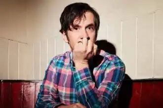 Man in plaid shirt looking pensive, sitting at a table against a wooden wall.