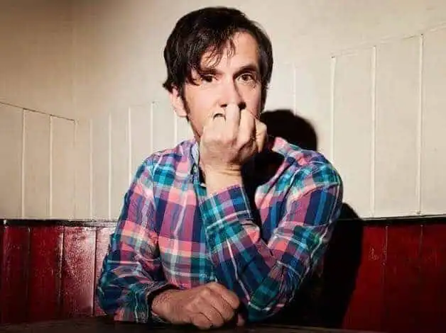 Man in plaid shirt looking pensive, sitting at a table against a wooden wall.