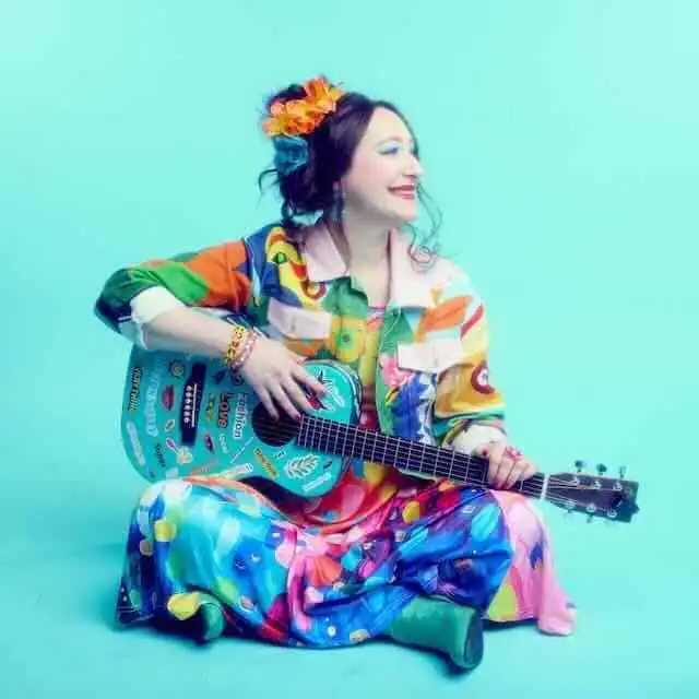 A joyful woman in colorful outfit playing guitar against a bright teal background, celebrating love and diversity.