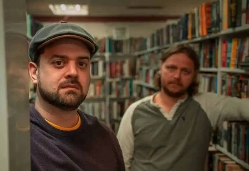 Two men standing in a lively bookstore aisle surrounded by colorful books.