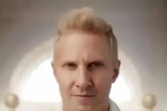 Close-up of a confident, serious man with blonde hair and blue eyes, wearing a white shirt.