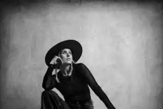 Black and white portrait of a poised woman wearing a wide-brim hat, stylish outfit, sitting against a plain background.