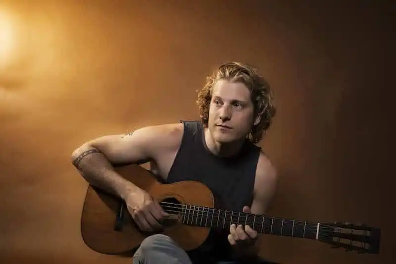 Alt text: Young man playing acoustic guitar with focused expression in warm lighting.
