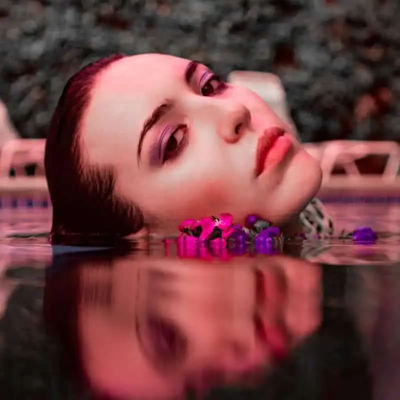 Beautiful woman relaxing in a pool with vibrant makeup, surrounded by colorful flowers.