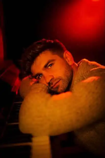 Close-up of a handsome man with dark hair and beard, resting his head on crossed arms, warm red and orange lighting.