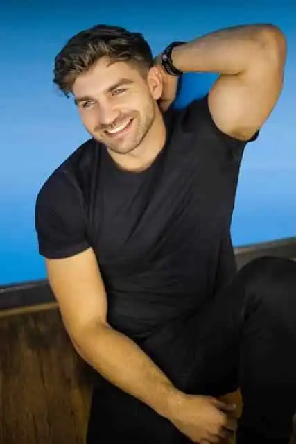 Smiling man with beard in casual black t-shirt, relaxed pose, blue background.