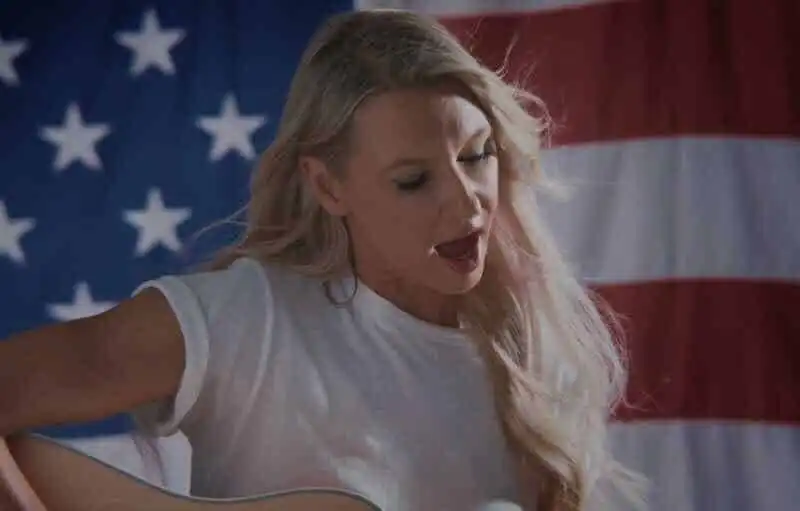 A woman with long blonde hair singing and playing guitar in front of an American flag backdrop, embodying patriotism and musical talent.