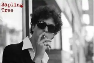 Black and white photo of a stylish person wearing sunglasses and a suit, making a shushing gesture.