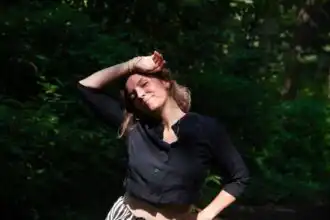 A woman in a black top smiling peacefully in a lush green outdoor setting.