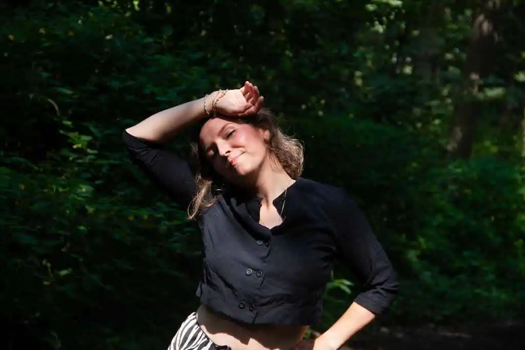 A woman in a black top smiling peacefully in a lush green outdoor setting.