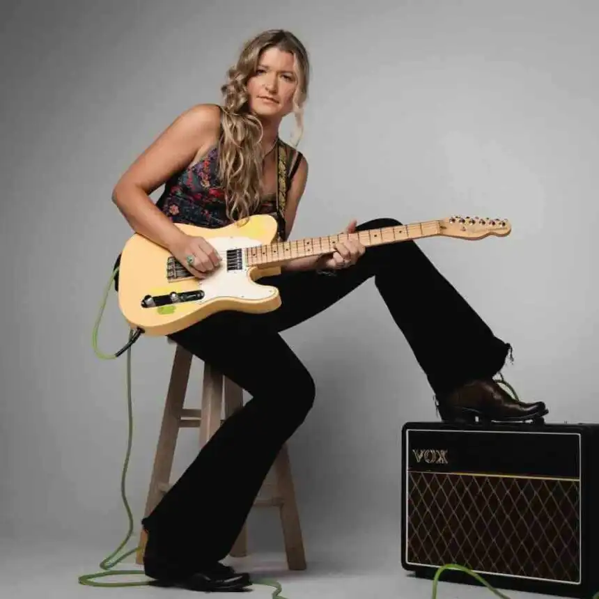 A woman with long wavy hair sitting on a wooden stool, playing an electric guitar with a Vox amplifier nearby.